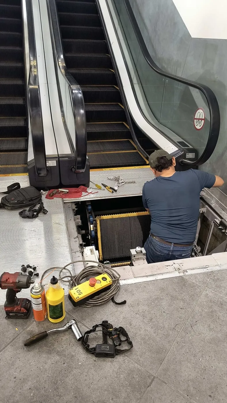 Técnico trabajando en la base de la escalera eléctrica, con herramienta y el control de maniobra en el piso.